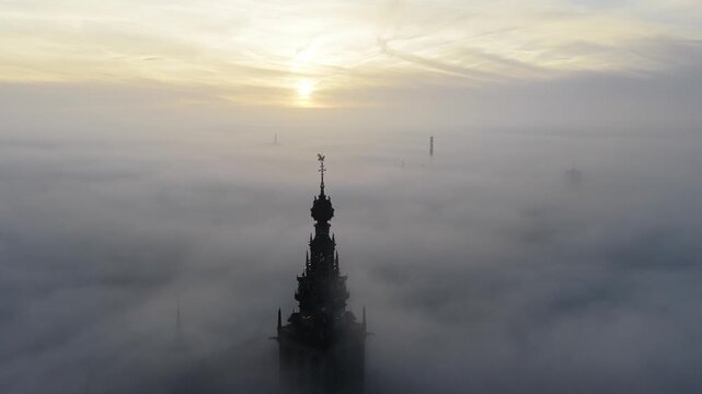 Aerial view of the Stevenskerk rising above Nijmegen during a foggy sunrise, with soft mist drifting through the historic streets and warm light coloring the sky. The church tower emerges from the haz