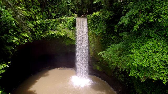 Tropical waterfall pouring into muddy pool amid dense rainforest at Tibumana Bali, drone dolly upstream