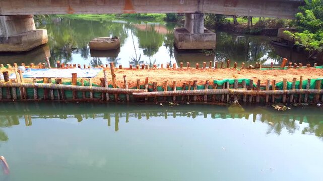 Aerial view of a temporary sand bund and wooden coffer dam under construction in a tropical river, featuring a concrete bridge and palm trees in the background. High-angle steady shot.