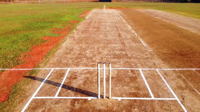 Aerial drone shot of a sunny cricket pitch, zooming out from the stumps to reveal the full field and boundary. Perfect for sports and outdoor recreation themes.