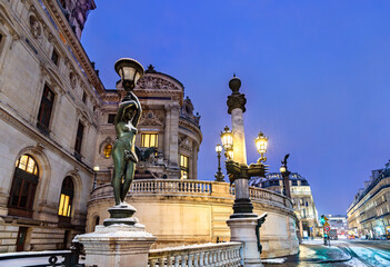 Obraz premium Palais Garnier is located in the 9th district of Paris, France. Winter scene features a bronze statue holding a lamp on the exterior facade covered in snow during the evening