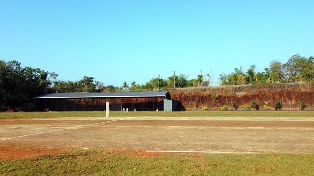 Wide drone shot of a quiet cricket ground and training nets under a clear blue sky, featuring a lush green field and a rustic red earth backdrop.