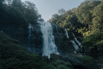 Fototapeta premium A majestic waterfall cascades down a mossy cliffside, surrounded by lush green vegetation and bathed in soft morning light.