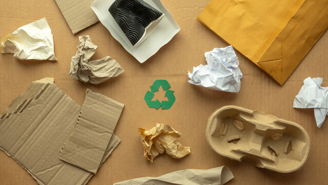 Flat lay of recyclable paper packaging waste with cardboard, envelopes, crumpled paper and green recycle symbol on brown background, eco friendly lifestyle, recycling and zero waste concept.