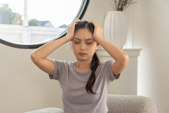 Young asian woman sitting on sofa feeling sick and touching head with headache migraine and pain suffering from stress vertigo and dizzy face expression