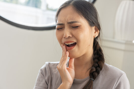 Sad asian woman suffering from strong toothache pain pressing cheek with hand inside home feeling unwell from dental problem and cavity needing dentist care while looking stressed with discomfort