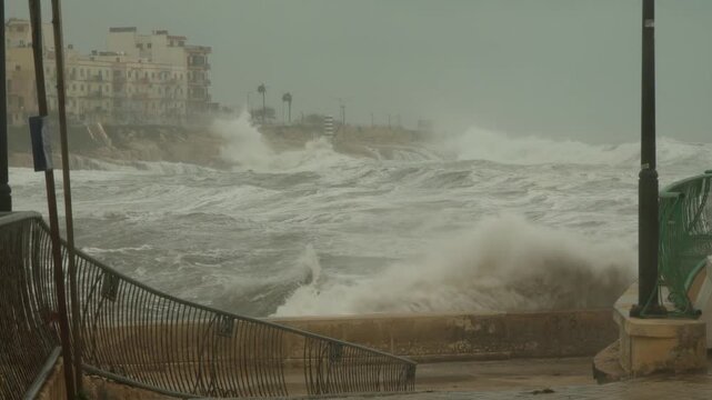 Slow-motion shot of a stormy scene in Marsaskala, Malta, during Storm Harry, captures rough seas crashing against a damaged promenade under overcast skies, showcasing extreme winter weather.