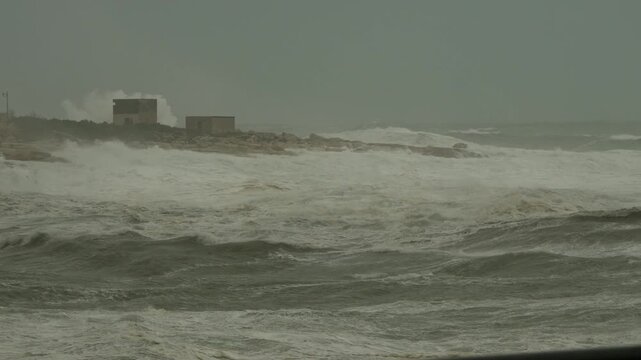 Slow-motion shot from the rough Mediterranean Sea during Storm Harry in Marsaskala, Malta, with powerful waves crashing against the rocky shoreline under overcast skies.