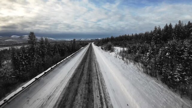 A snow-covered two-lane road stretches through a forested mountain landscape under a cloudy sky.
