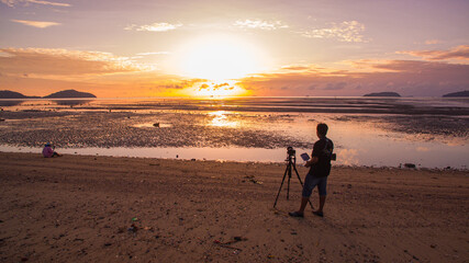 A lone photographer stands on a quiet beach with a tripod, capturing a golden sunrise over shallow tidal waters. The expansive sky, reflections on wet sand, and distant islands create a calm  © Narong Niemhom