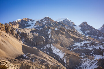 Obraz premium Breathtaking view of a rugged Himalayan mountain range featuring snow-dusted slopes, sharp rocky peaks, and layered alpine terrain beneath a clear blue sky. The vast, untamed landscape
