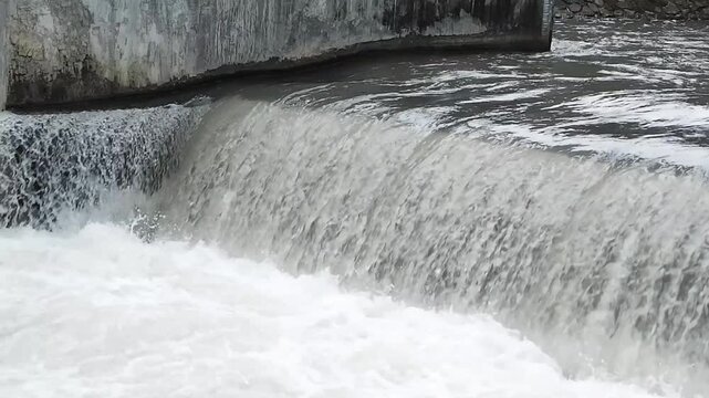 Fast-flowing river water cascades over a small concrete weir, creating white foam and dynamic currents, illustrating water management, natural motion, and rural infrastructure.