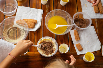 Child hands mixing tiramisu cream in plastic bowl, top view of dessert preparation at kids cooking workshop on wooden table
