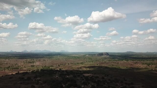 Wide aerial landscape of northern Mozambique (Nampula region), with dry savanna, woodland stretching to the horizon and isolated inselberg hills rising from the plain beneath scattered cumulus clouds.