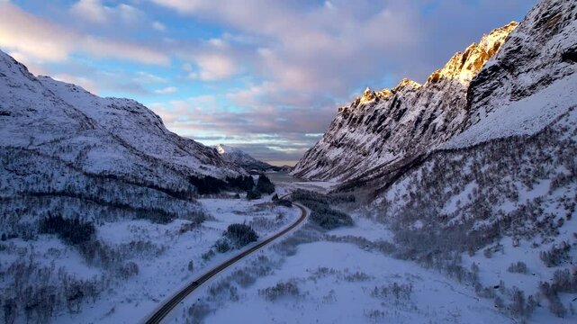 4K vertical rise drone shot revealing the iconic E10 Surrounded by frozen fjords and steep snow capped peaks in the Arctic Lofoten islands at sunrise.