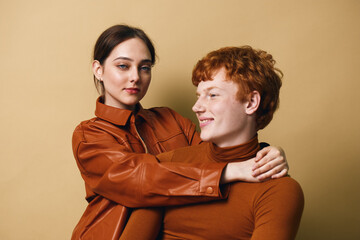 Obraz premium Young couple with brown hair and casual outfits embracing each other on beige background. Woman with leather jacket holding smiling redhead man in close pose.