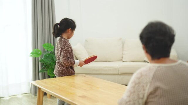 Young girl practicing table tennis with elderly woman in cozy indoor setting, highlighting family bonding and intergenerational playtime in living room