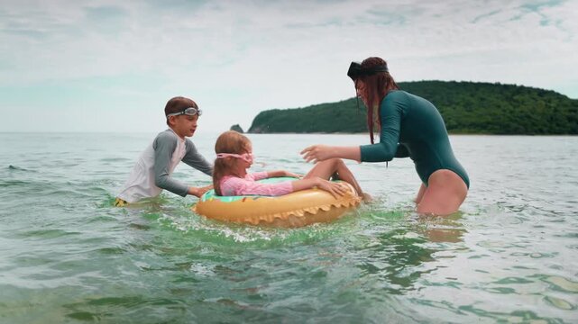 Cheerful siblings and mom play together with inflatable float in warm sea. Smiling faces and active movement reflect joy, fun and carefree emotions of family beach leisure time.