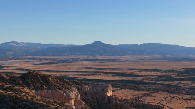 Drone moves sideways showing white sandstone cliffs in the foreground with Cerro Pedernal in the distance. The high desert landscape features eroded sedimentary rock and volcanic basalt peaks.