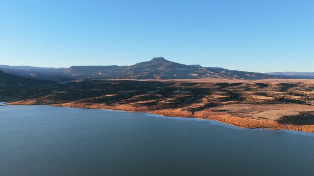 A peninsula of arid land extends into the deep blue Abiquiu Lake reservoir water below Cerro Pedernal. Late afternoon sun hits the volcanic high desert plateau of northern New Mexico state.