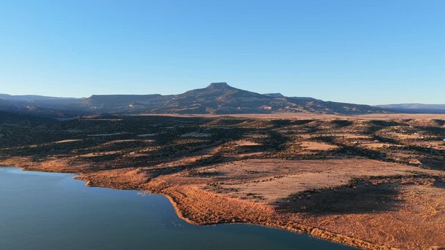 Drone view highlights the prominent volcanic basalt mesa of Cerro Pedernal rising above the calm water of Abiquiu Lake. The remote high desert landscape features arid brush and expansive plateau topog