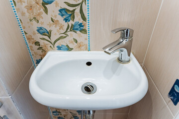 Fototapeta premium Ceramic Sink Details. Compact White Ceramic Sink With Chrome Tap In Tiled Corner Bathroom. Small Bathroom Fixture With Ceramic Basin And Floral Accent Tiles In Apartment Renovation