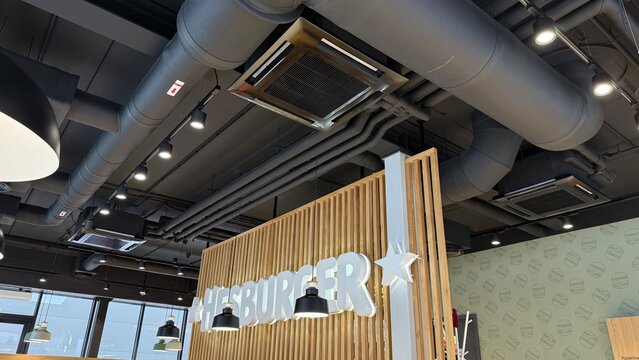 Fast food restaurant interior with exposed industrial ceiling HVAC ventilation ductwork cassette air conditioning units and pendant lighting above Hesburger brand signage. 7 February 2026, Lithuania