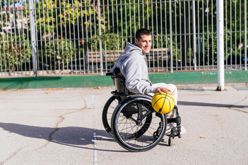 Obraz premium Young man with a prosthetic leg is sitting in a sports wheelchair, holding a basketball on an outdoor court
