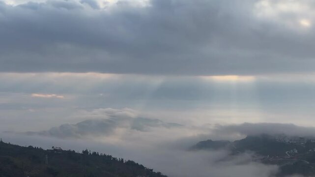 A cinematic 1080p aerial timelapse featuring a dramatic mountain landscape at dawn. The footage captures golden sunbeams (crepuscular rays) filtering through heavy, fast-moving clouds and thick valley