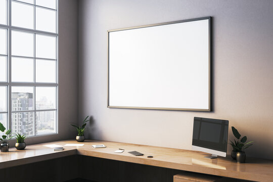 Blank canvas frame on office wall above modern wooden desk setup with computer and plants in light-filled corner room interior design scene. 3D Rendering
