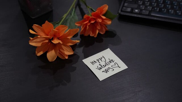 Close-up of hand placing &ldquo;Happy Valentine Day&rdquo; sticky note on dark office desk beside orange flowers and computer keyboard, soft indoor lighting.