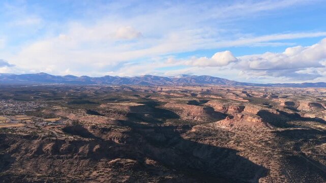 A wide aerial vista captures the expansive volcanic mesas and deep canyons of the Pajarito Plateau. Snow-capped mountain peaks sit on the horizon beneath a bright blue sky in northern New Mexico.