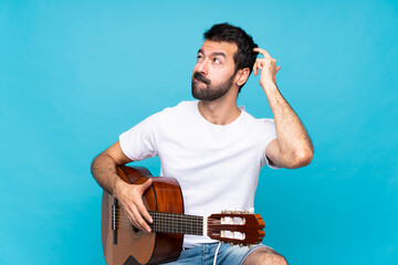 Young man with guitar over isolated blue background having doubts while scratching head