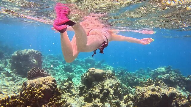 Female swimmer with pink aqua socks glides through clear blue water above coral reef, showcasing underwater movement and vibrant marine environment. Snorkeling on vacation.