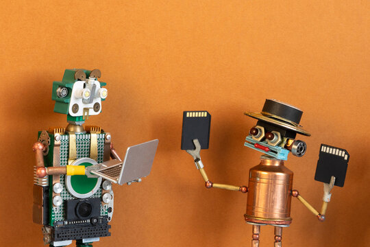 Happy smiling toy robot holding memory cards showing them to another robot with open laptop. Data transfer concept, digital collaboration poster