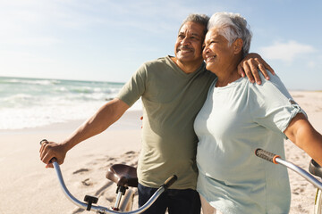 Obraz premium Happy senior multiracial couple with bicycles looking away at beach against sky on sunny day
