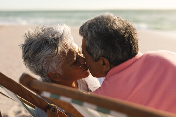 Obraz premium Affectionate senior multiracial couple kissing while sitting on folding chairs at beach