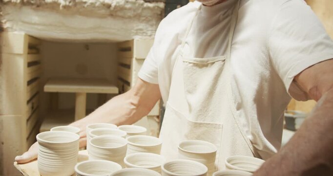 Smiling senior caucasian man wearing apron taking fired pots from kiln at pottery workshop