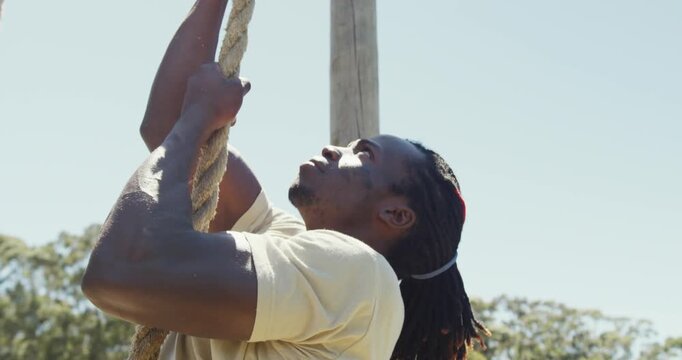 Fit african american male soldier with deadlocks climbing rope on obstacle course in sun