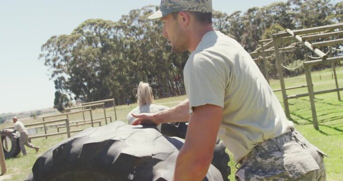 Fit caucasian male soldier in cap rolling tractor tyre on obstacle course in the sun