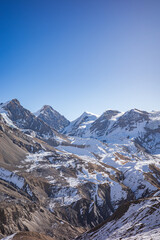 Panoramic view of a snow-capped mountain range with deep alpine valleys, rugged rocky slopes, and layered winter terrain beneath a clear blue sky. The vast scale, natural contrast, and sense of remote