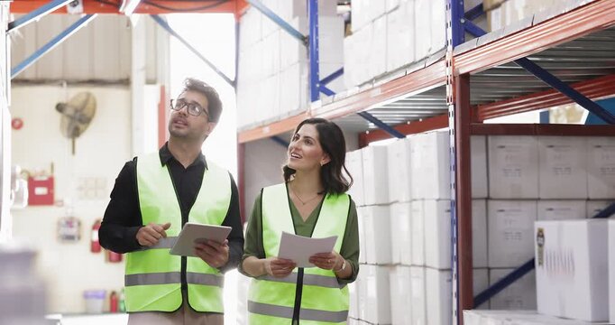Male Hispanic warehouse manager reporting to female boss, walking along metal shelves, pointing at boxes, showing storage. Mentor training interns to male inventory, count goods, check waybills