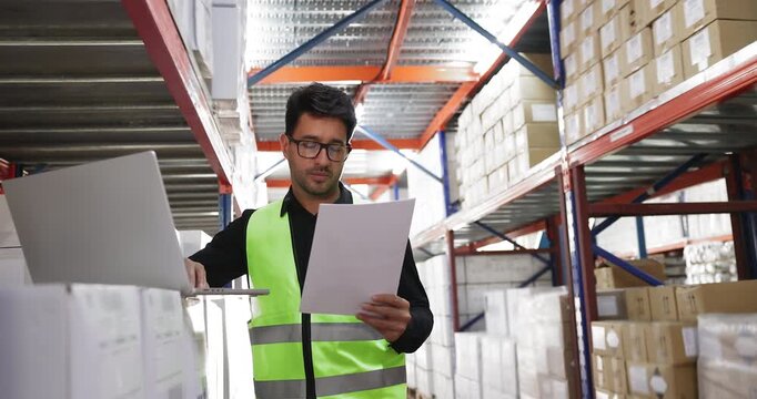 Hispanic warehouse employee man checking paper waybill, using laptop computer in storage space, entering part numbers after delivery, typing, consulting paper report