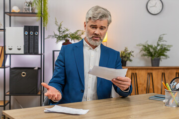 Naklejka premium Middle-aged businessman at home office stacks unpaid bills feeling pressure then angrily throws them aside. Freelancer guy at table strategizes expense cuts seeking relief from mounting urgent debt.