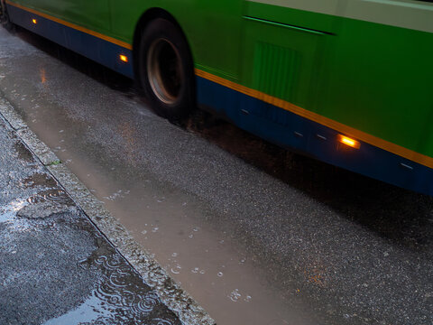 A public transport bus splashes through water on a rainy day, creating ripples and reflections on the asphalt surface