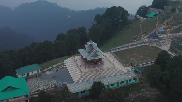 Dynamic slow drone pullout showcases Hatu Mata Temple's tiered roofs, plaza, and green auxiliary buildings, expanding to reveal thick surrounding forests and misty mountain layers.