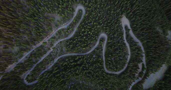 Winding mountain switchbacks form flowing patterns as a narrow road snakes through dense forest and steep slopes, aerial top down descend