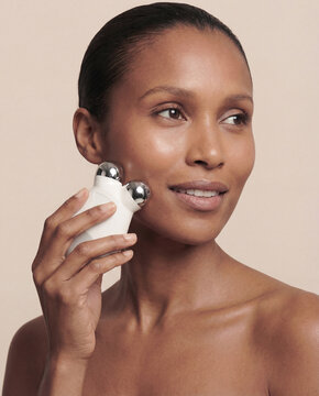 Woman using microcurrent facial roller on cheek, studio skincare portrait on beige background
