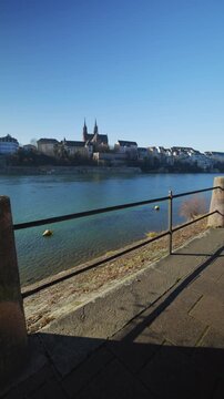 Vertical static high-angle shot from a stone terrace overlooking the Rhine river toward the historic Basel Minster (Basler M&uuml;nster) with its twin towers and the old town skyline under a clear blue sky