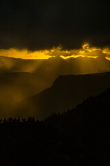 Dramatic mountain landscape with layered ridges and golden sun rays breaking through storm clouds, creating atmospheric light and deep shadows. © Stoca